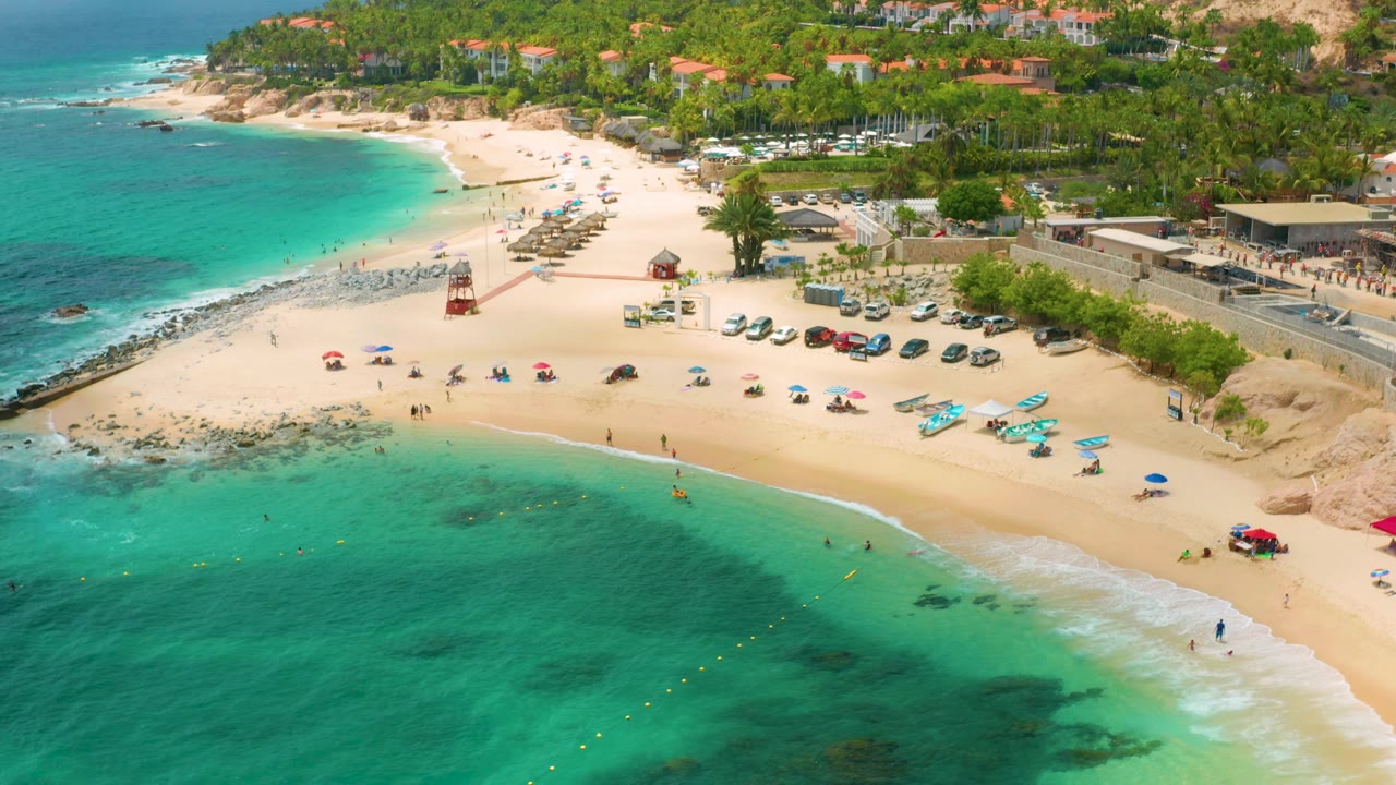 Aerial view over Palmilla Beach Cabo with shallow turquoise water across the lower frame, pale sand with umbrellas and beach craft, parked cars near the access road, and palms and buildings beyond.