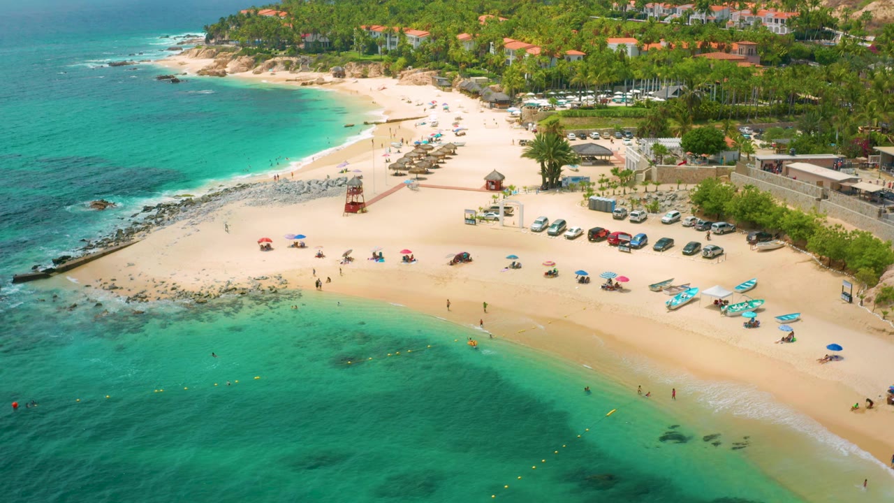 A second aerial frame of Palmilla Beach Cabo, Mexico, shows the shoreline slightly farther along, with shallow green-blue water, beach umbrellas, parked cars, palm trees, hillside buildings, and distant people on the sand.