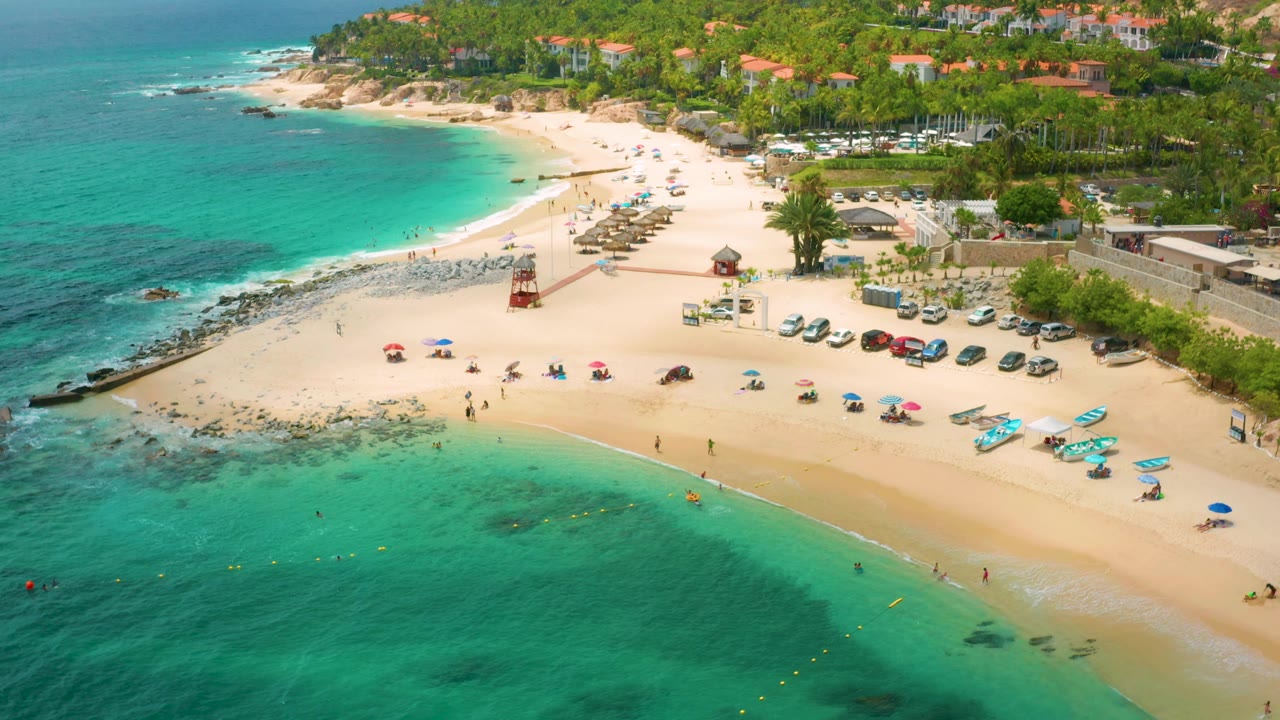 Aerial view at Palmilla Beach Cabo, Mexico, with the waterline crossing the lower frame, umbrellas and small craft on the sand, parked cars beside palms, hillside buildings in back, and distant beachgoers visible.