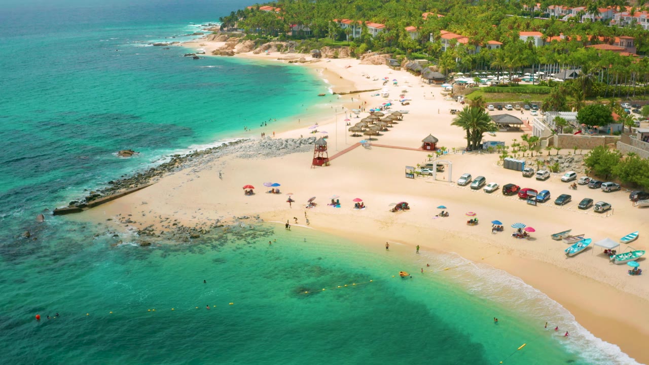 High aerial beach view with swimmers, umbrellas, parked cars, palm trees, and clear green-blue water.