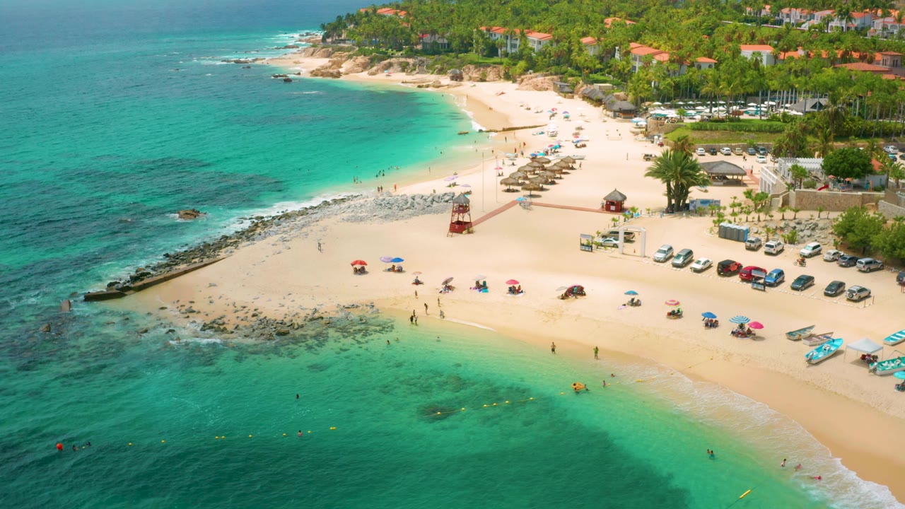 Palmilla Beach Cabo, Mexico appears from above with the shoreline curving across the frame, swimmers near the shallows, beach umbrellas below, and developed resort grounds behind the palms.