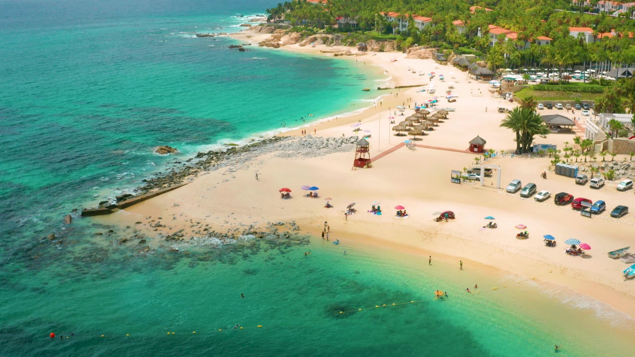 Aerial view of Palmilla Beach Cabo, Mexico, with turquoise shallows in the foreground, people near the waterline, colorful umbrellas on sand, and palm-lined buildings beyond the beach.
