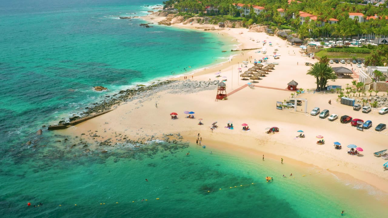 At 0:02, the Palmilla Beach Cabo shoreline curves across frame with dark reef patches under turquoise water, scattered beachgoers, umbrellas along the lower sand, and a lifeguard tower near center right.