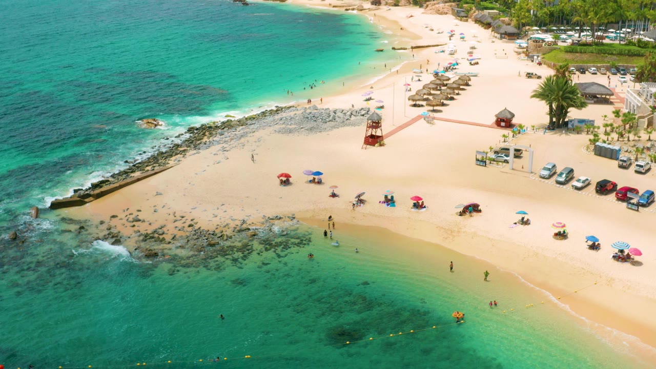 Aerial frame over Palmilla Beach Cabo, Mexico, showing turquoise water meeting pale sand, scattered umbrellas, rocky shoreline, parked cars, palms, and small distant beachgoers.
