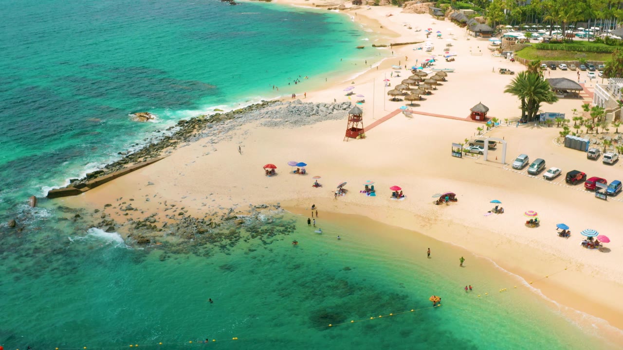 Aerial frame over Palmilla Beach Cabo, Mexico, with clear green-blue water in the foreground, a curved sandy beach, umbrella clusters, a red tower, and tiny visitors along the waterline.