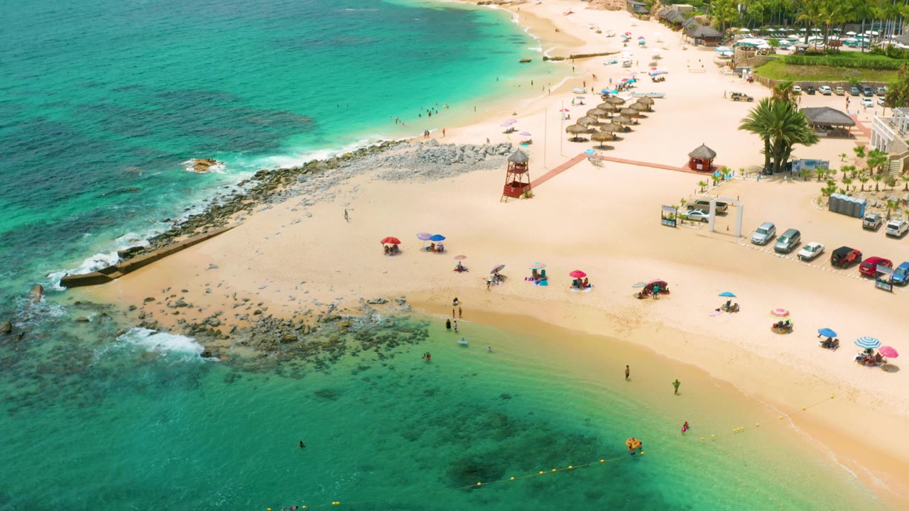 Aerial frame over Palmilla Beach Cabo, Mexico, showing a wider sweep of turquoise bay water, pale beach, straw shade structures, umbrellas, palms, parked cars, and distant people on the sand.