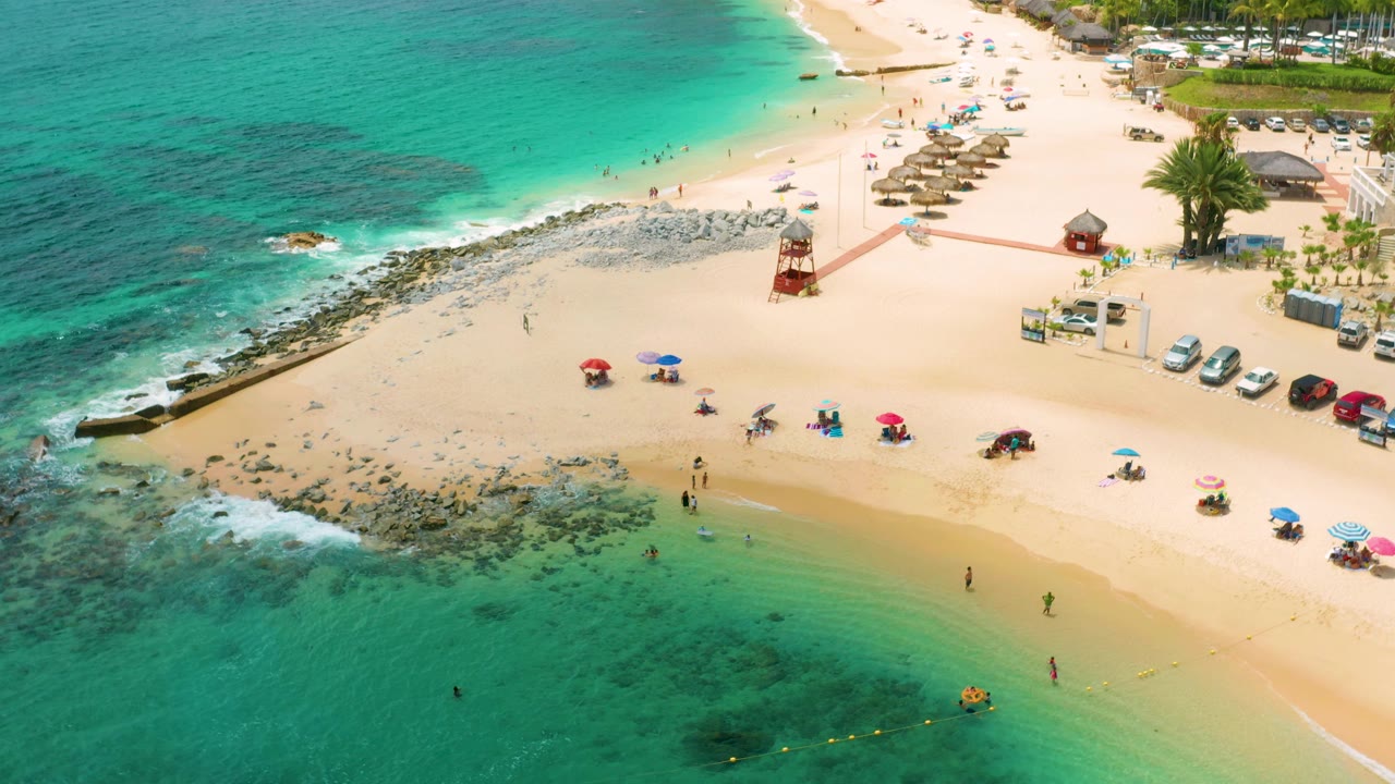 Drone view of beach umbrellas, shallow water, rocks, and resort edge.