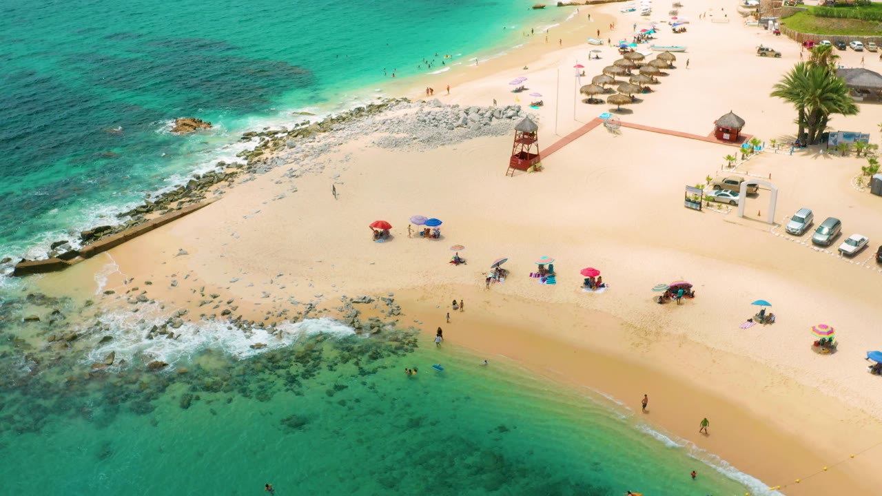 Overhead beach scene with waves, umbrellas, rocks, a tower, and people on the sand.