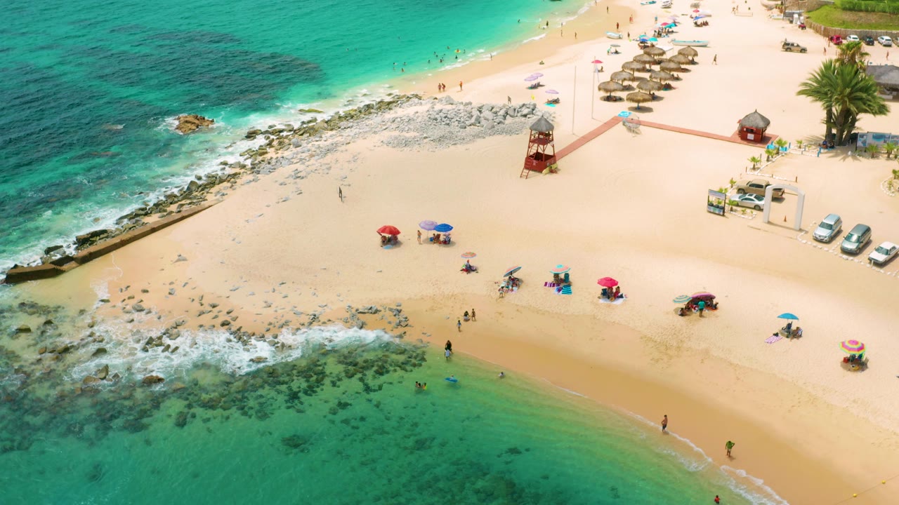 Aerial view over Palmilla Beach Cabo with clear green-blue water at the bottom, a broad sand band, umbrellas set apart from each other, and palms and vehicles near the upper right.
