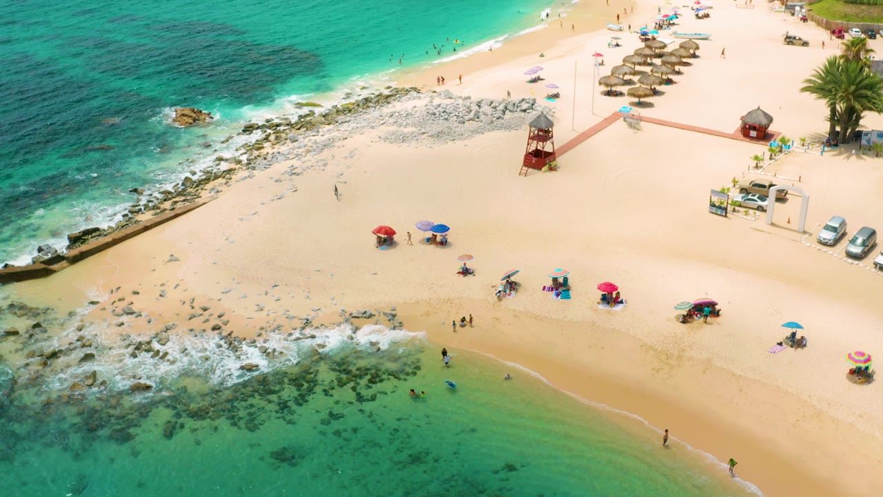 Aerial shoreline with waves washing over rocks and beach umbrellas on pale sand.