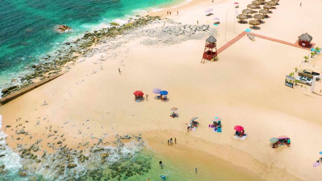Overhead view of Palmilla Beach Cabo, Mexico with turquoise surf at left, pale sand, scattered umbrellas, a red lifeguard tower, and small visible beachgoers.