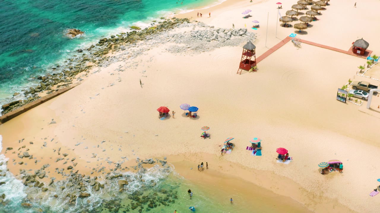 Aerial beach view with shoreline rocks, open sand, umbrellas, and small beachgoers.