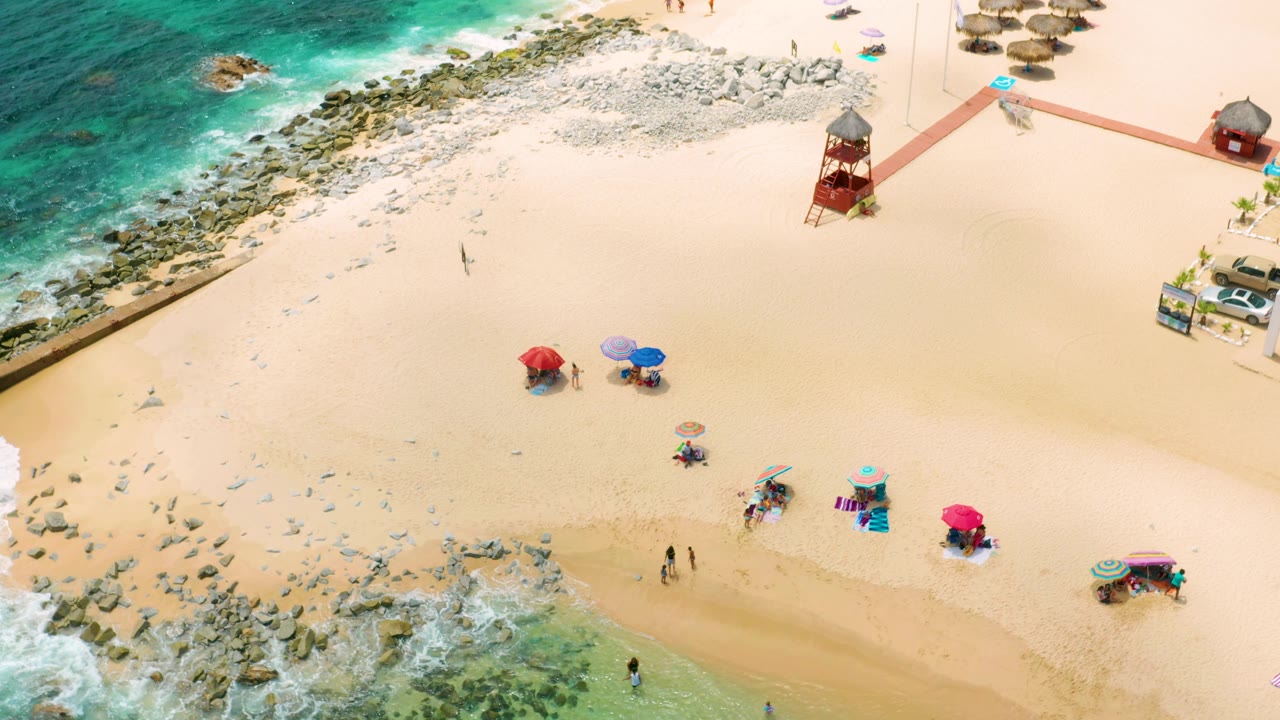 High aerial view of Palmilla Beach Cabo, Mexico with the red lifeguard tower near the upper sand, rock barriers beside the surf, and several umbrellas with small visible people below.