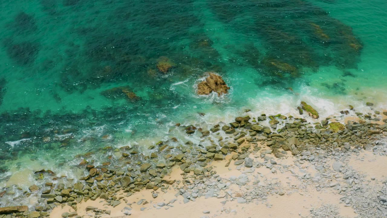 Aerial frame from Palmilla Beach Cabo, Mexico, showing green-blue sea water over submerged rocks, a small tan rock near center, foamy wash, pale beach sand, and no visible people.