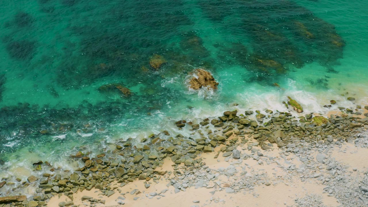 Overhead view at Palmilla Beach Cabo, Mexico, with darker submerged rock patches in turquoise water, a foamy band over shoreline stones, pale sand below, and no visible people.