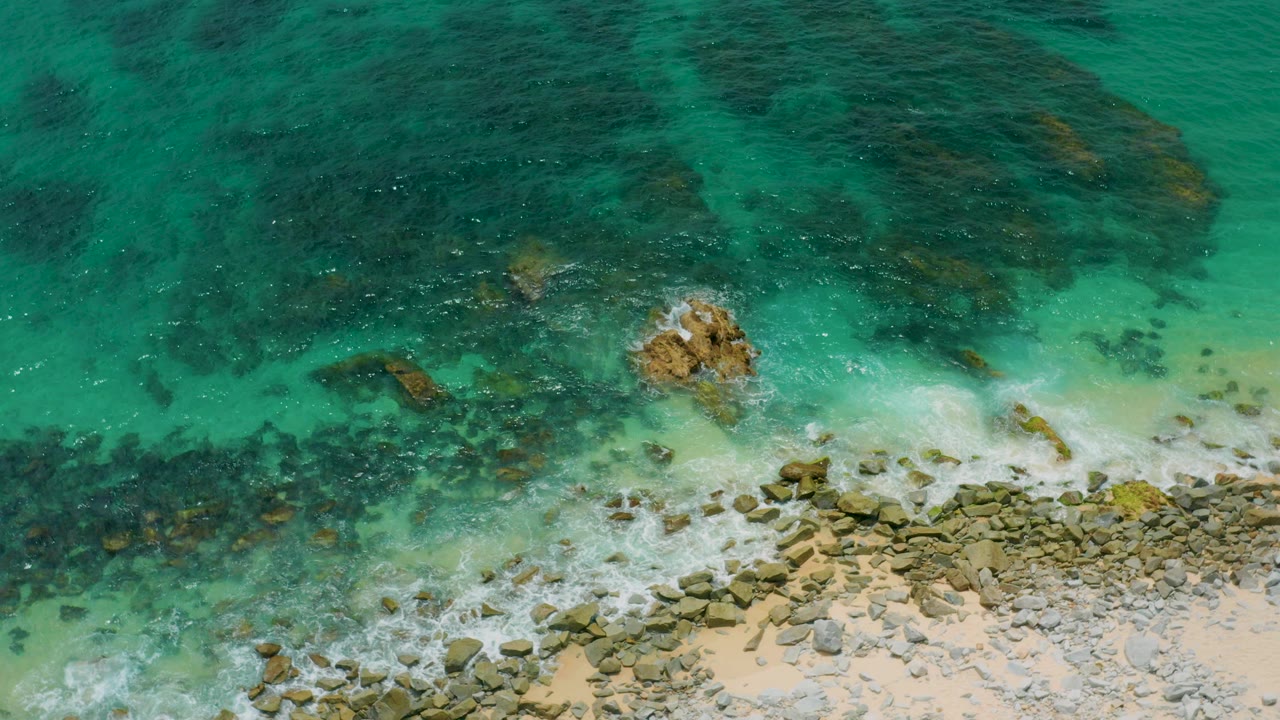 Overhead view at Palmilla Beach Cabo, Mexico, showing green water over dark submerged rocks, a tan rocky shore at the bottom, small surf, and no visible people.