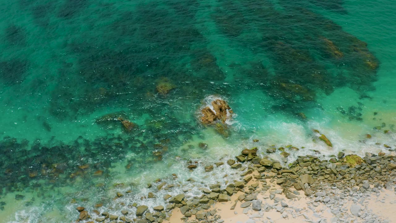 Aerial frame over Palmilla Beach Cabo, Mexico, with a small exposed rock surrounded by white water, dark submerged rocks across the upper frame, and no visible people.