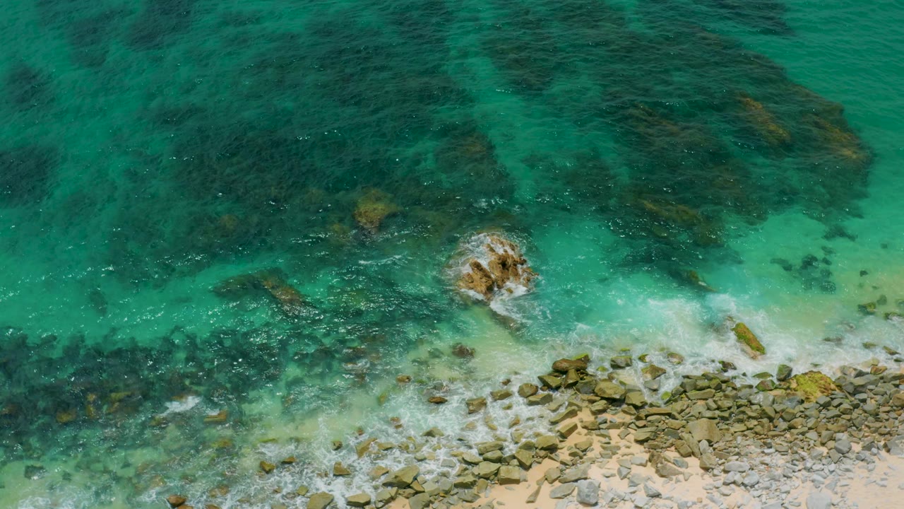 Overhead shoreline frame from Palmilla Beach Cabo, with white surf spreading over the lower rocks, green water in the center, dark underwater patches above, and no visible people.