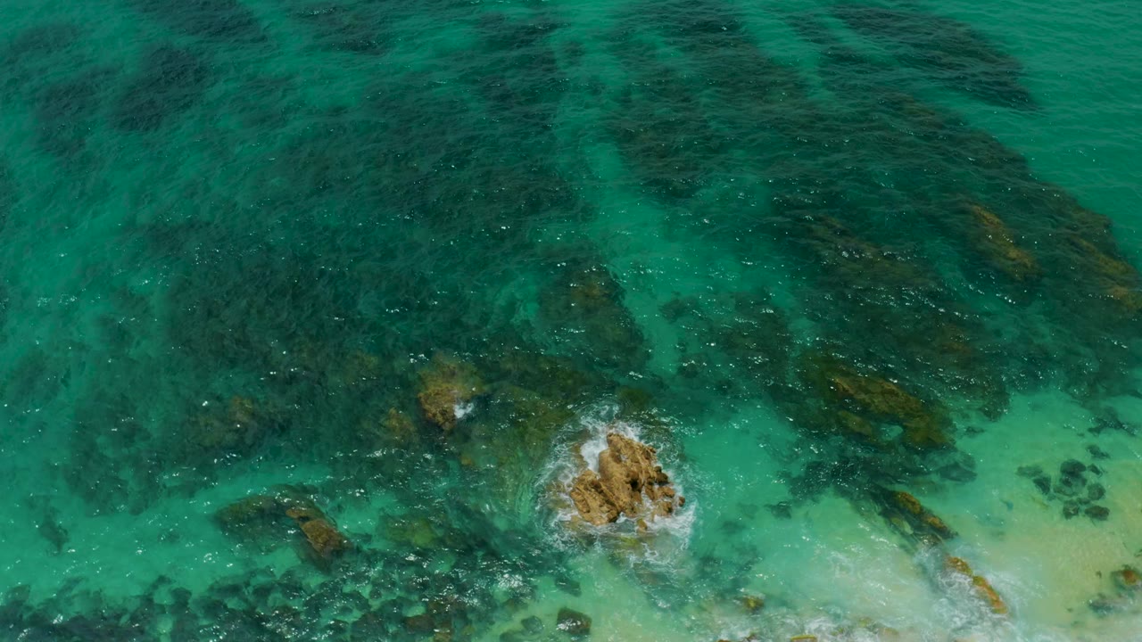 Overhead Palmilla Beach Cabo water view with the exposed rock shifted lower in frame, dark underwater formations spreading across the top, pale sand to the right, and no visible people.