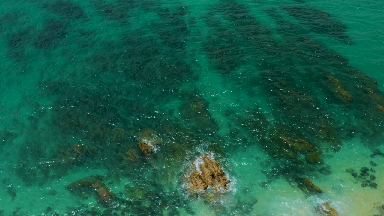 Top-down view at Palmilla Beach Cabo, Mexico, showing reef-darkened water across the upper frame, a foaming rock near the lower right, pale shallows, and no visible people.