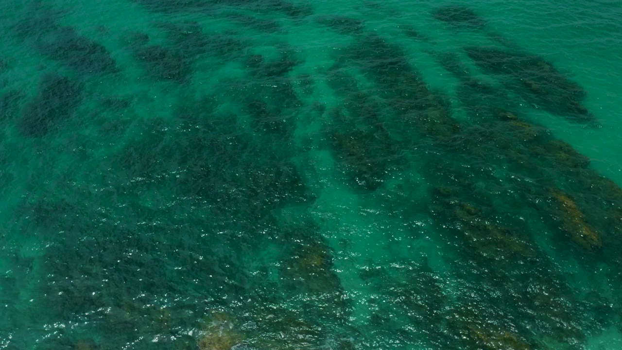 Aquamarine water at Palmilla Beach Cabo, Mexico, fills the frame with broad dark seafloor patches under a lightly rippled surface, no visible people.