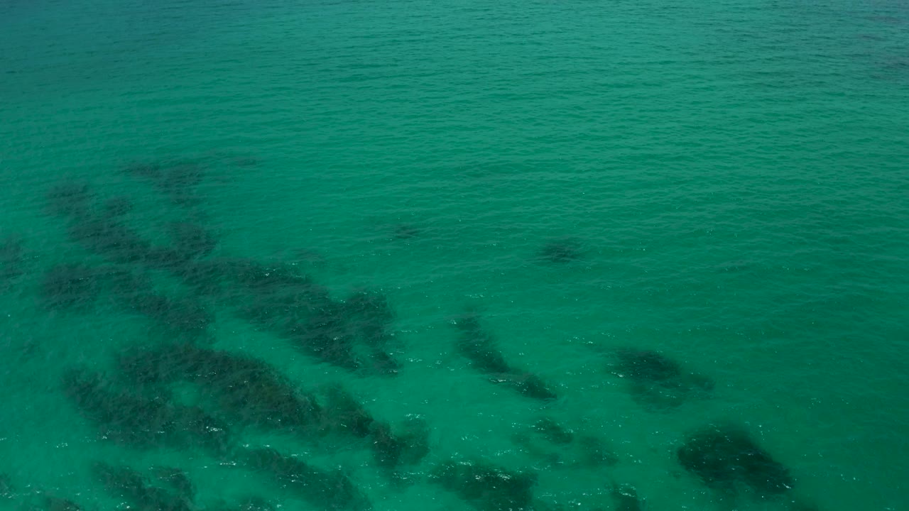Clear green ocean at Palmilla Beach Cabo, Mexico, seen from above with larger dark reef-like shapes toward the right side and no visible people.