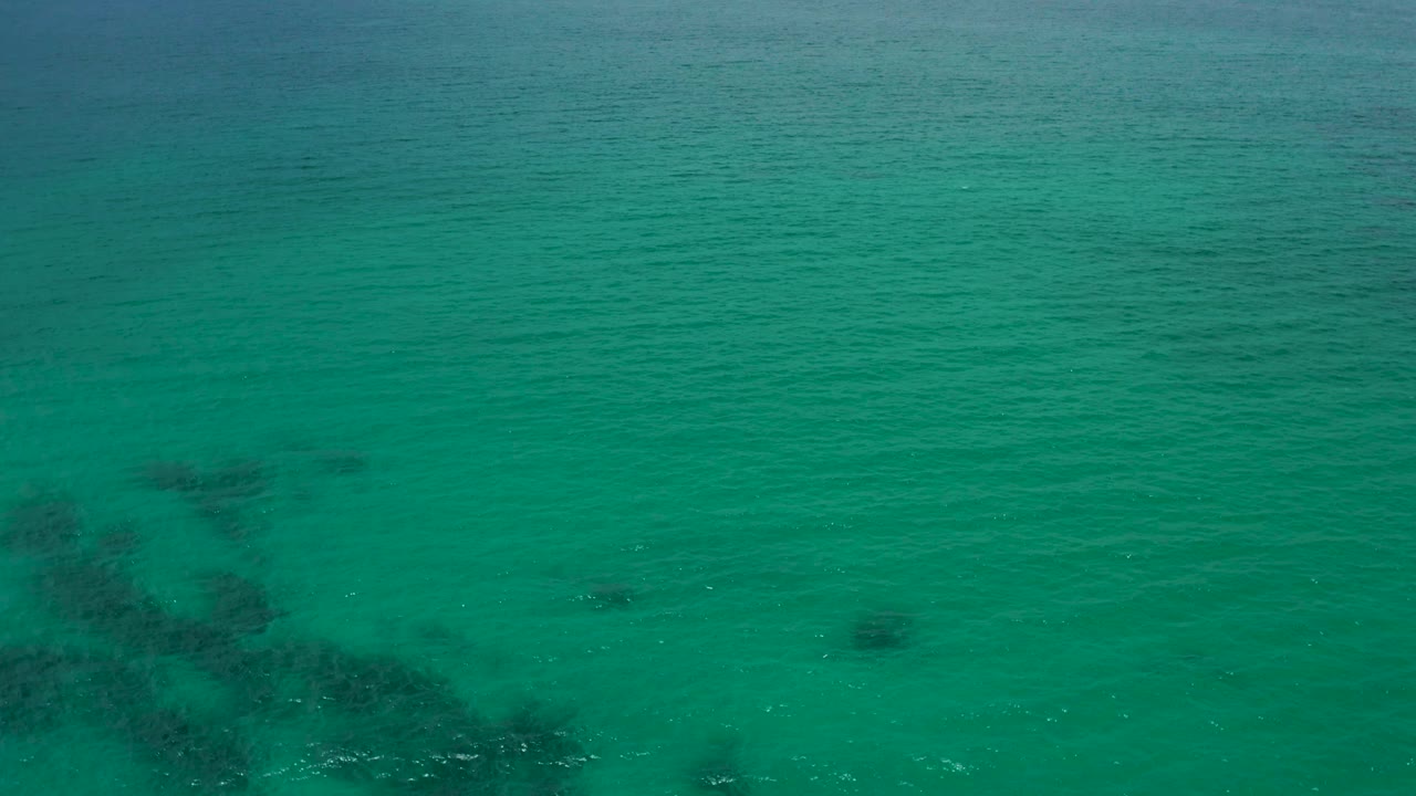 Aerial frame over Palmilla Beach Cabo, Mexico showing open green water, faint ripples, and several darker submerged areas with no visible people.