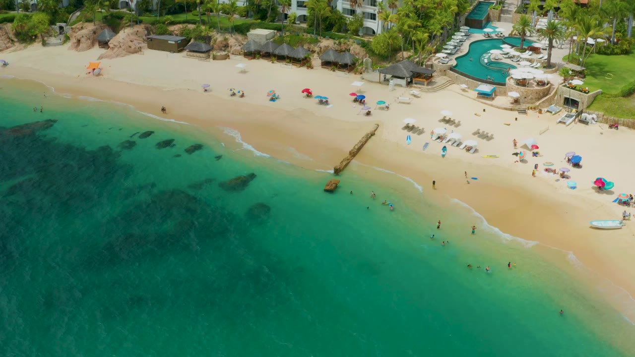 High aerial view over Palmilla Beach Cabo, Mexico, with turquoise water at left, a sandy beach with visible people, colorful umbrellas, lounge chairs, a narrow pier, and resort pools near the top edge.
