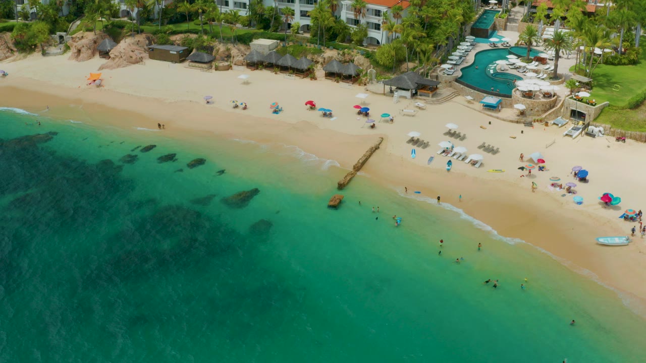 Aerial frame over Palmilla Beach Cabo, Mexico showing turquoise water at lower left, pale sand with small beach umbrellas and beachgoers, a narrow jetty, a pool terrace, palms, and resort buildings above the beach.