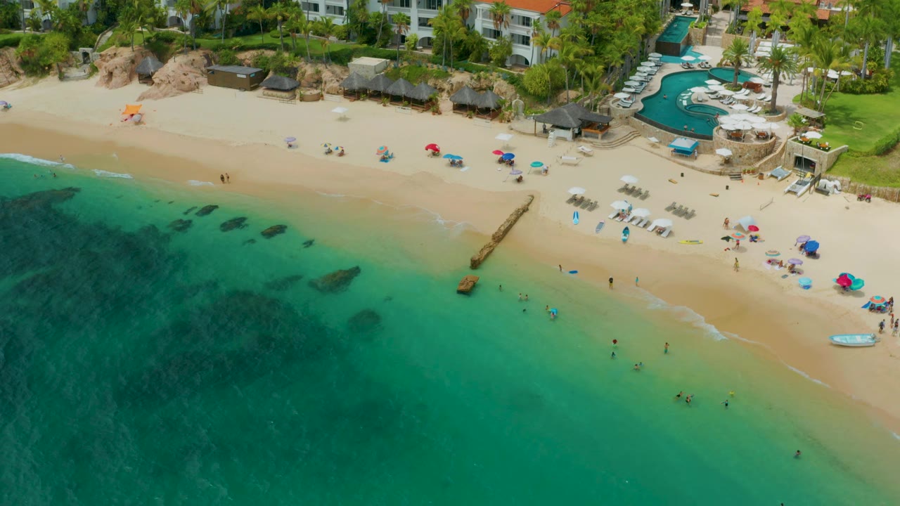 Drone-height frame over Palmilla Beach Cabo, Mexico showing the shoreline running diagonally, small figures in the surf, colorful umbrellas on the sand, a small white boat near the right edge, palms, pools, and red-roofed resort buildings.