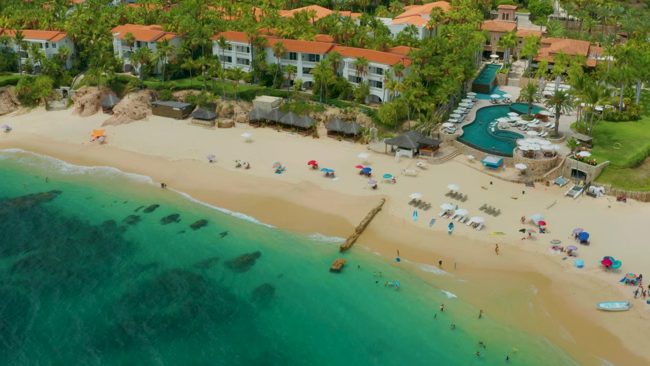 A high-angle view over Palmilla Beach Cabo, Mexico shows turquoise water in the foreground, beach umbrellas and lounge chairs on the sand, a curved pool, palm trees, and red-roofed shorefront buildings with tiny distant people visible.