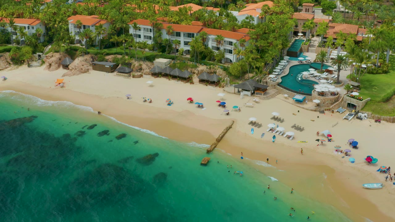 At 0:01, Palmilla Beach Cabo, Mexico appears from above with more open water in the foreground, pale surf along the sand, scattered umbrellas, a pool terrace, palms, and tiny distant beachgoers visible.