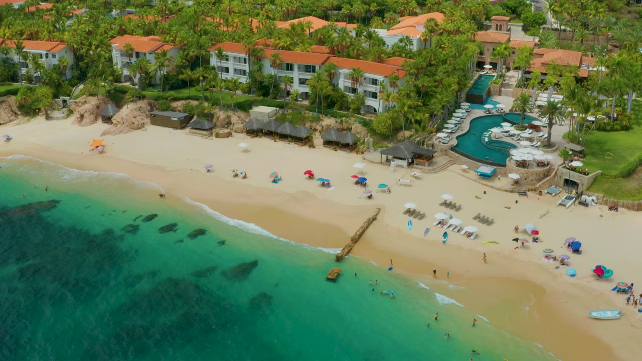 At 0:02, the aerial frame shows Palmilla Beach Cabo, Mexico with darker patches beneath turquoise water, a narrow surf line, beach umbrellas, a curved pool area, palm trees, and tiny distant people visible on the beach and in the water.