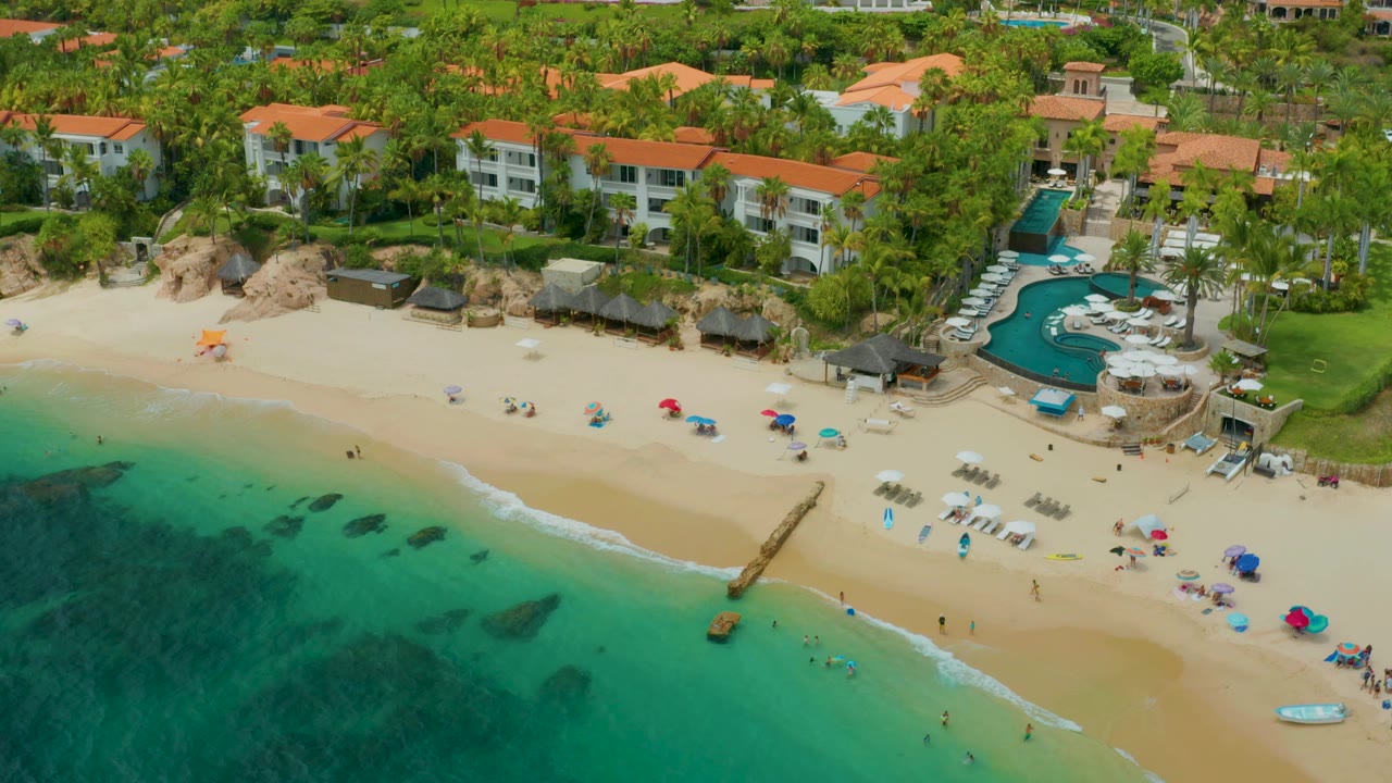 High aerial view of beach, shallow water, umbrellas, pool, palms, and shorefront buildings.
