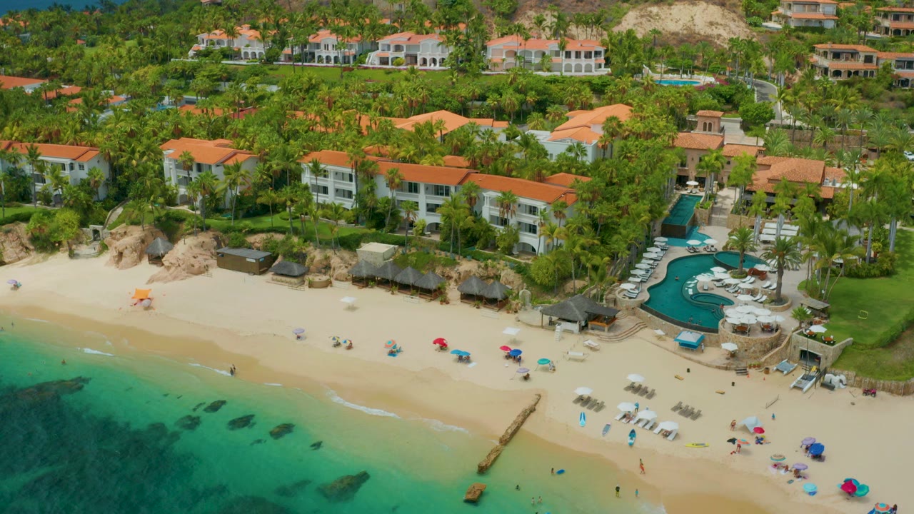 Aerial frame over Palmilla Beach Cabo, Mexico, showing turquoise water at lower left, pale sand, beach umbrellas, palm landscaping, red-tile-roof buildings, and no clearly visible people.