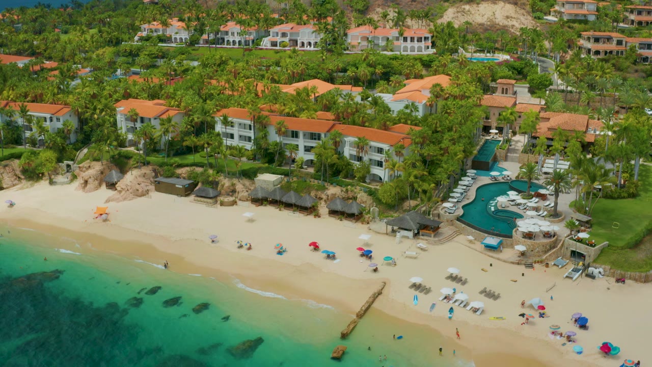 Aerial view at Palmilla Beach Cabo, Mexico, with blue-green ocean along the lower edge, a broad sandy beach, a curved pool area beside palms, red-roof buildings, and no clearly visible people.