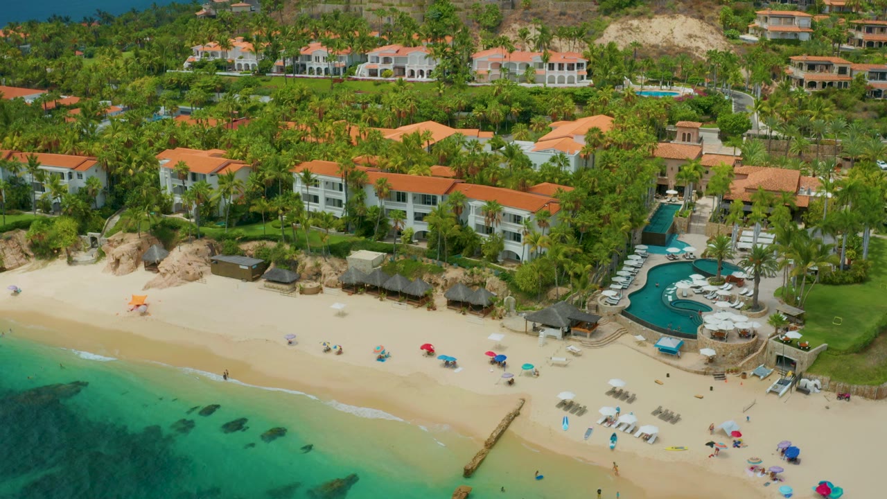High aerial frame from Palmilla Beach Cabo, Mexico, showing shallow turquoise water, a pale beach with scattered umbrellas and chairs, dense palms, resort buildings, hillside homes, and no clearly visible people.