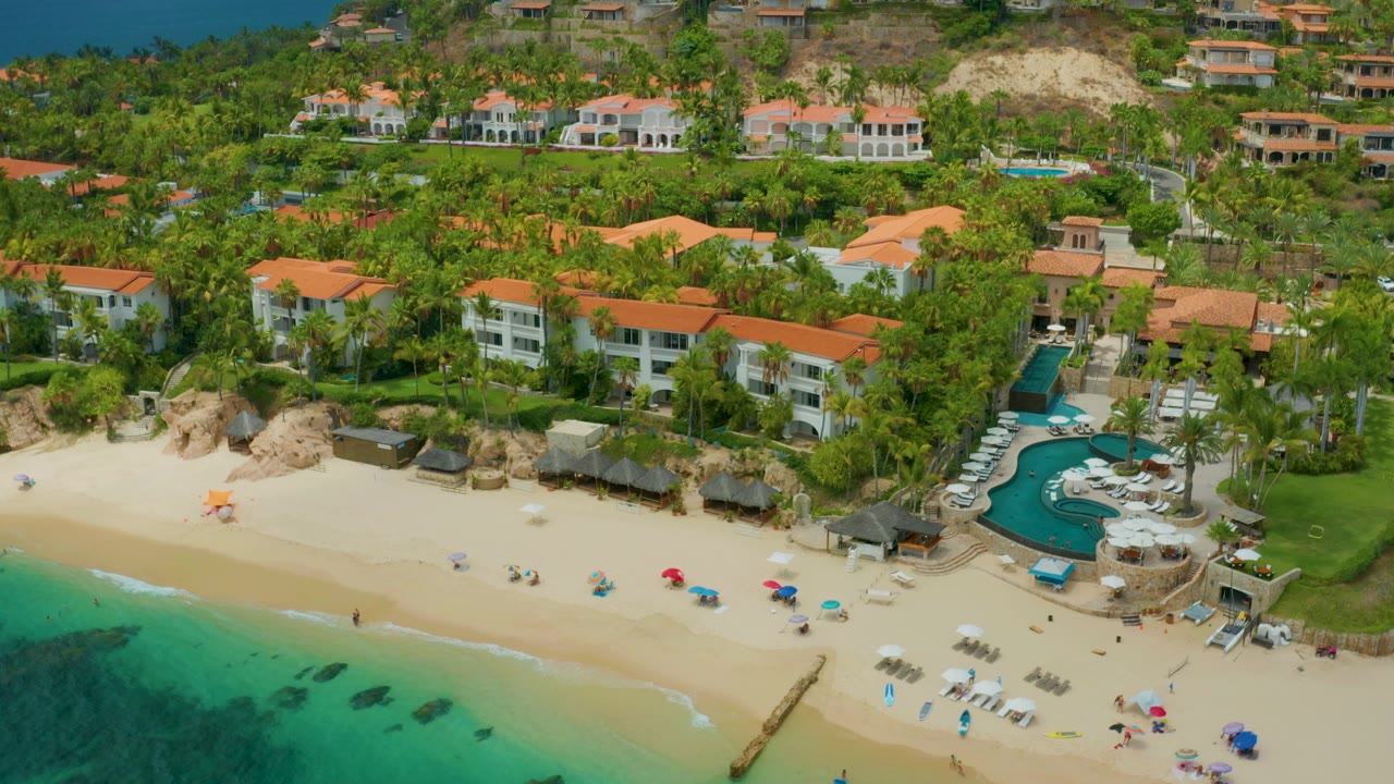Wide aerial view of beach umbrellas, calm water, palms, and resort buildings.