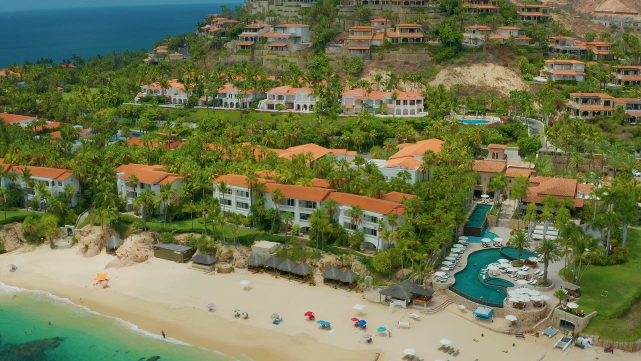 Opening aerial frame over Palmilla Beach Cabo, Mexico with red-roof resort buildings, a curved pool, pale sand, turquoise water, hillside homes, and no visible people.