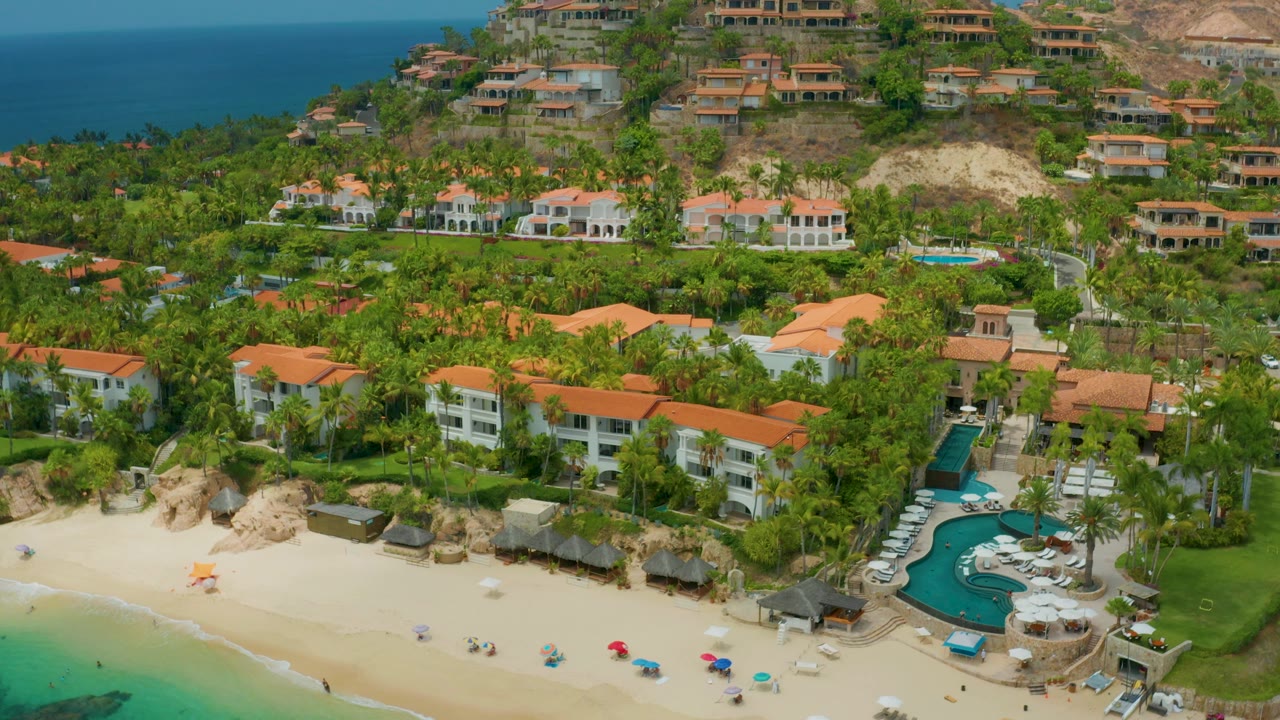 Wide Palmilla Beach Cabo frame showing resort buildings and palms below hillside homes, with sandy beach and blue-green water at the bottom and no visible people.