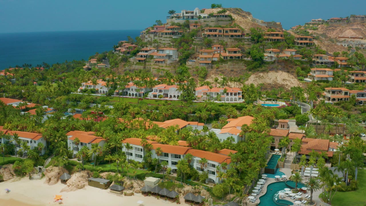 Opening aerial frame over Palmilla Beach Cabo, Mexico, with terracotta roofs, green palms, a sandy beach edge, hillside homes, blue ocean on the left, and no visible people.
