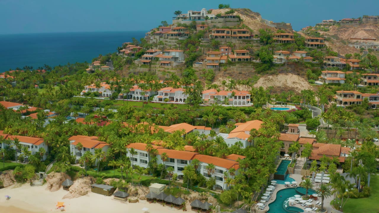 Aerial view one second into the Palmilla Beach Cabo clip, showing the same resort complex with the ocean band at left, hillside houses above, pools below, and no visible people.