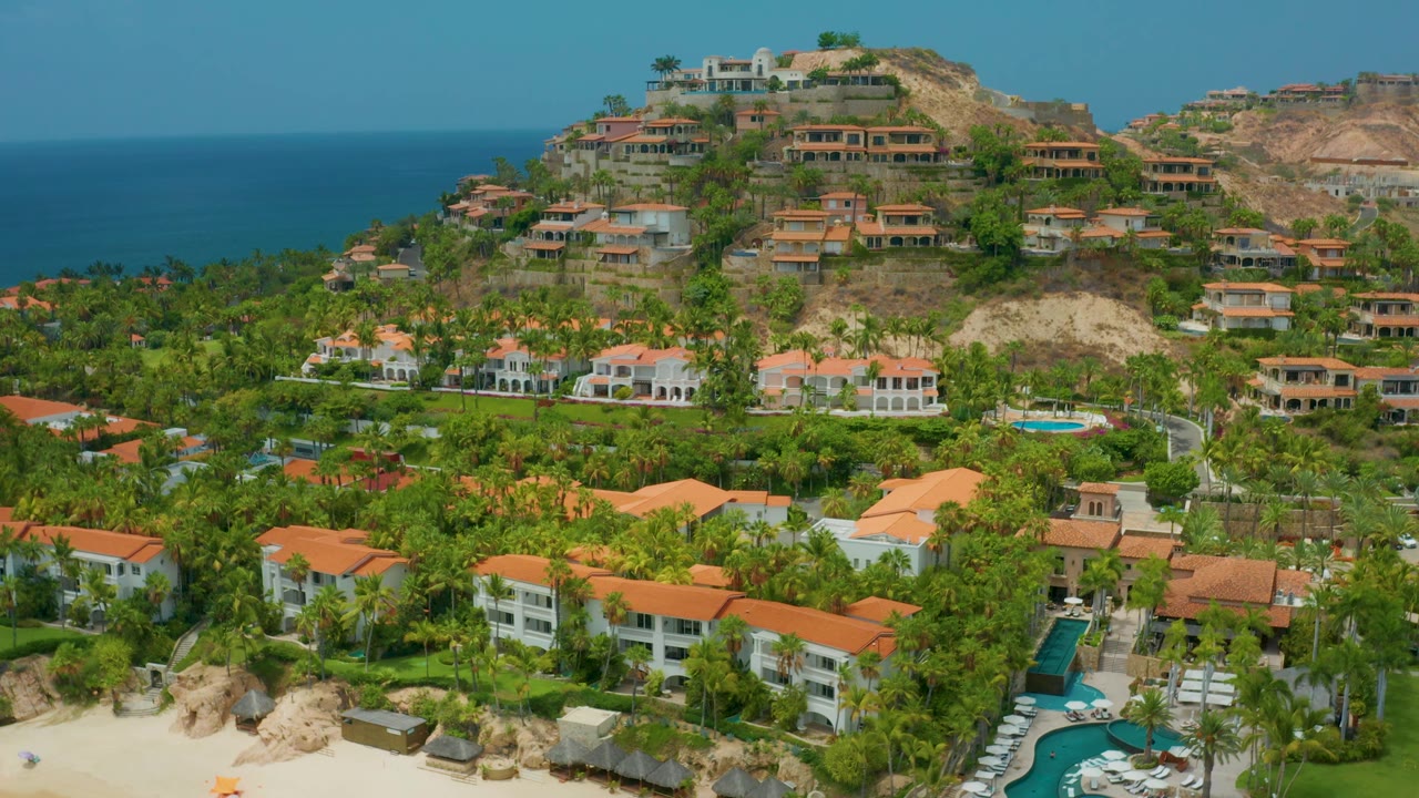 Palmilla Beach Cabo aerial frame with beach and pool areas along the lower edge, layered palm trees and orange roofs across the middle, dry hillside homes behind, and no visible people.