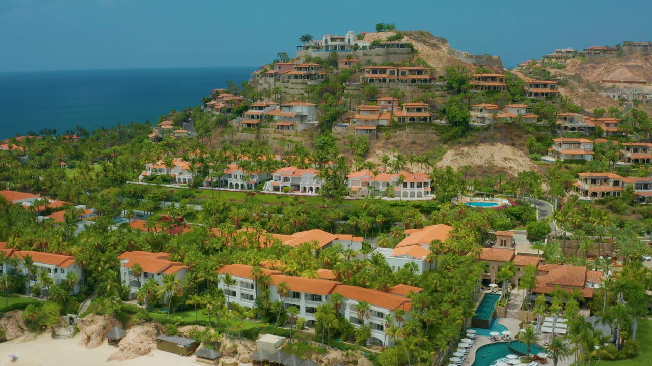 Aerial resort landscape with ocean horizon, palms, and hillside residences.