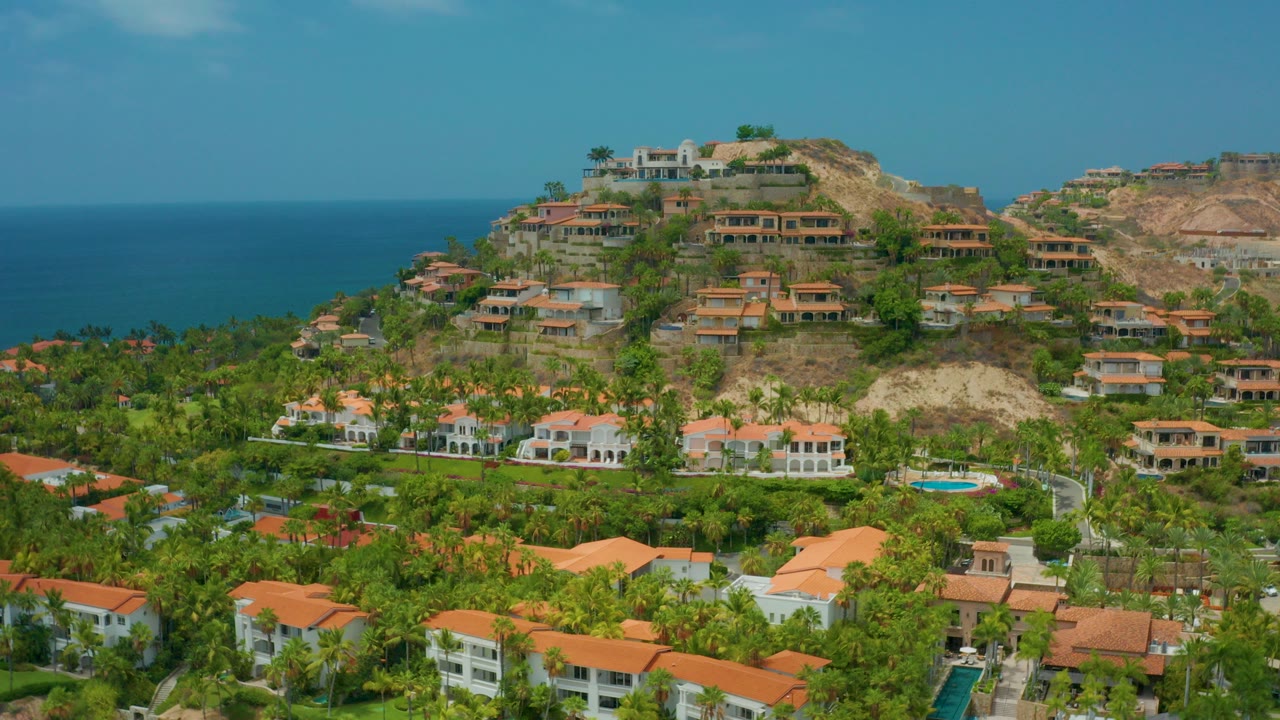Palmilla Beach Cabo, Mexico aerial frame with terracotta-roofed homes in green palms, hillside residences above, blue ocean behind, and no visible people.