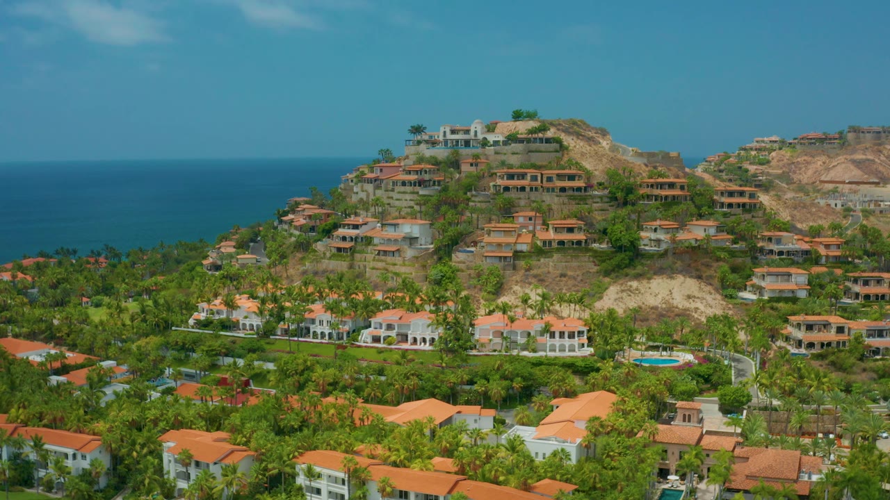 Aerial coastal town view with orange roofs, green palms, hill, and ocean