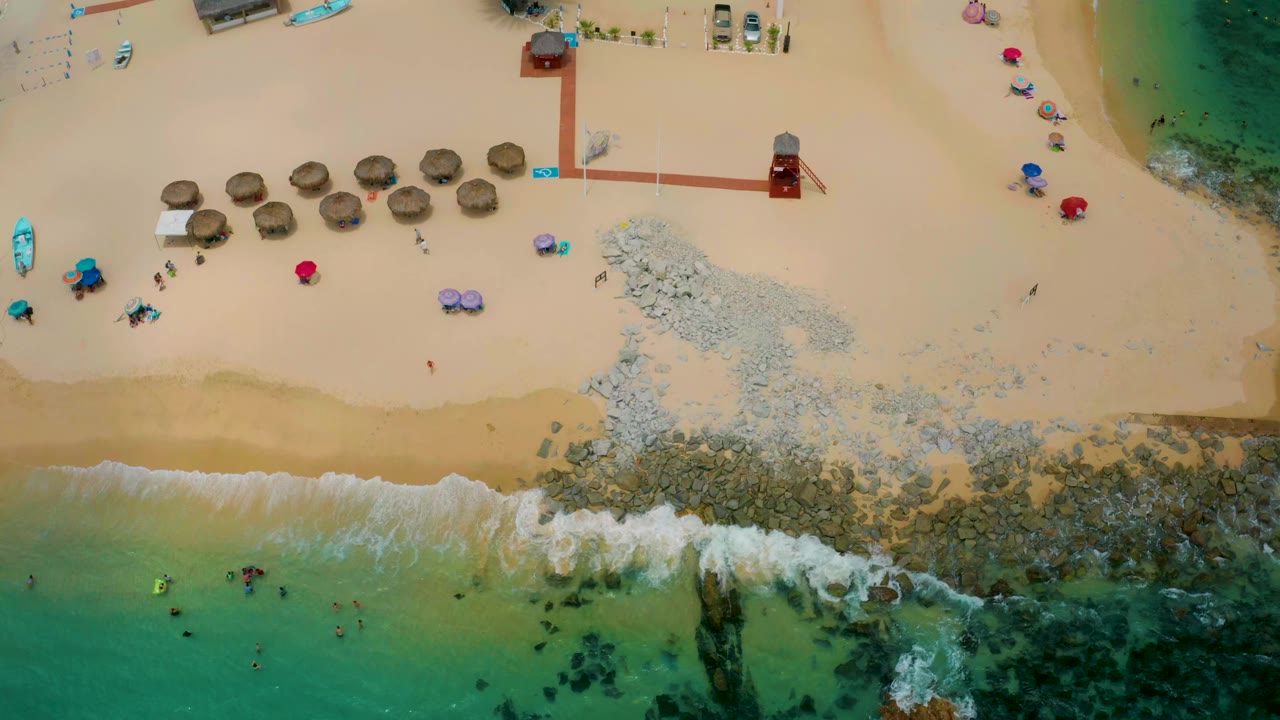 Aerial beach view with palapas, rocky shoreline, swimmers, and shallow green water.