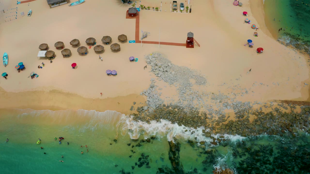 Top-down view of Palmilla Beach Cabo with the rocky section and breaking surf lower right, palapa huts across the sand, colorful umbrellas near the edge, and distant people visible as small figures.