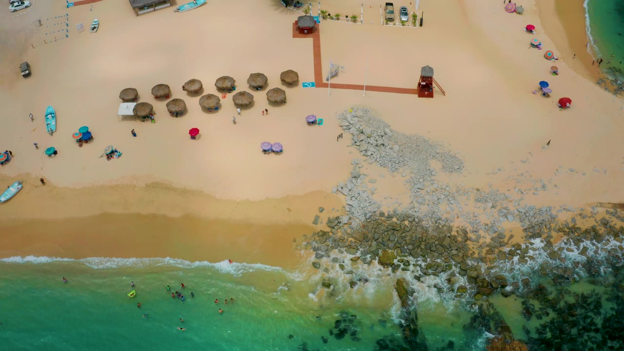 Aerial frame over Palmilla Beach Cabo, Mexico, showing the wave line lower in frame, rocks spreading across the center, and a row of thatched umbrellas on the upper sand.