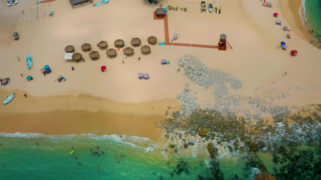 Top-down view of Palmilla Beach Cabo, Mexico, with swimmers as small figures in the water, a sandy strip, central rocks, and thatched shade umbrellas near the top.